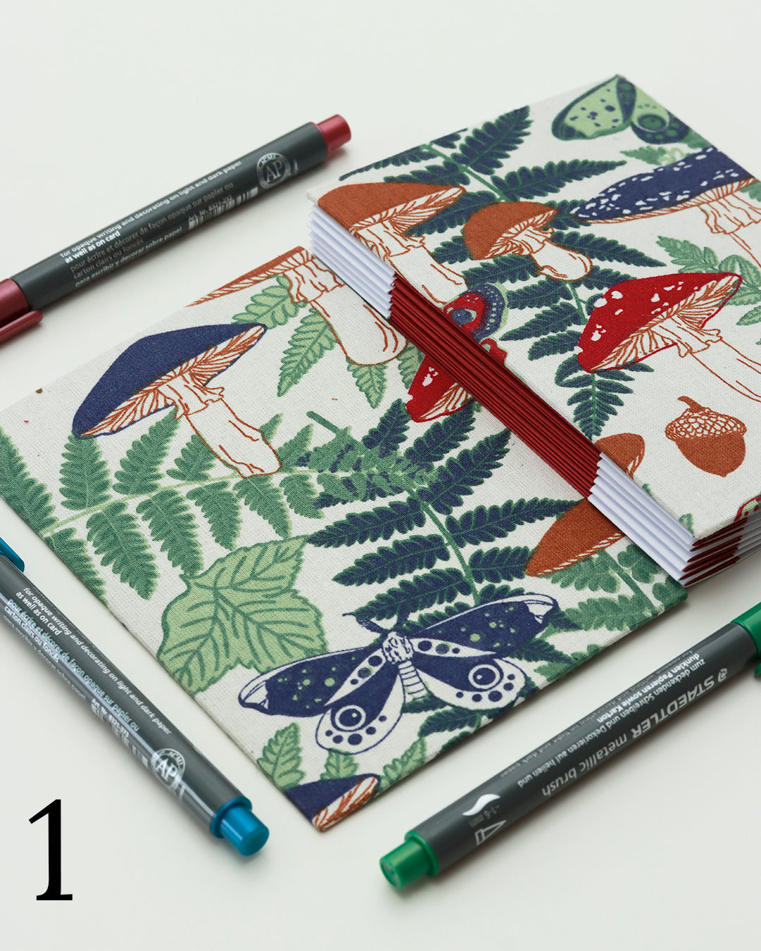 Quaderno per hand lettering con rilegatura australiana, copertina in tessuto con stampa di funghi, felci e ghiande nei toni di verde, rosso e blu su fondo panna, dorso a fisarmonica rosso.