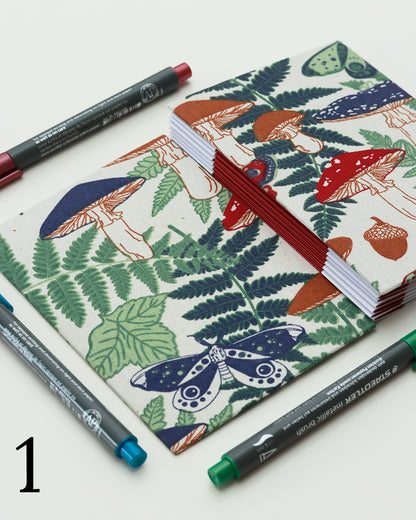 Quaderno per hand lettering con rilegatura australiana, copertina in tessuto con stampa di funghi, felci e ghiande nei toni di verde, rosso e blu su fondo panna, dorso a fisarmonica rosso.