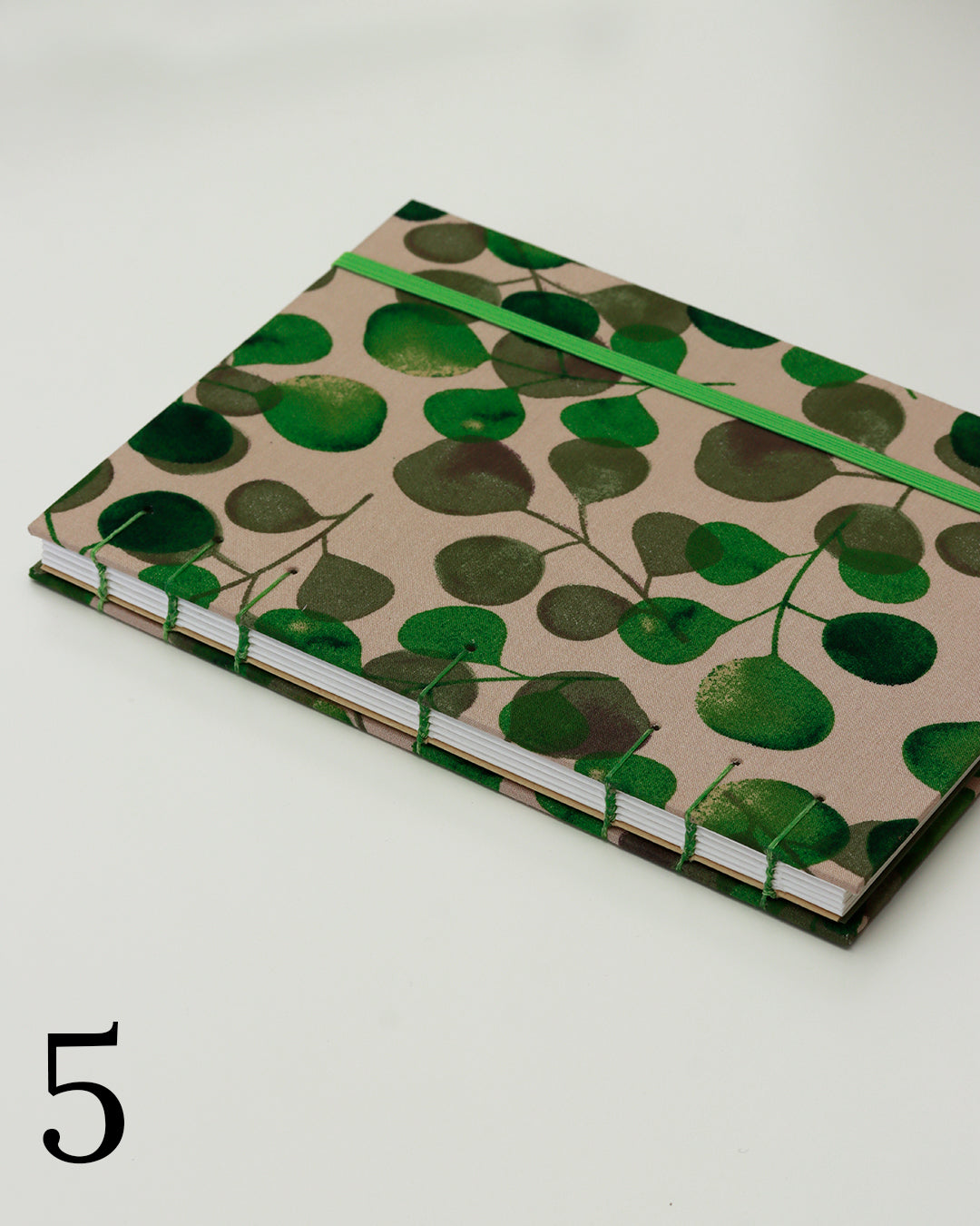 Quaderno per calligrafia con rilegatura copta, copertina in tessuto con foglie di eucalipto verdi su fondo tortora chiaro, elastico e filo verde, occhielli grigi.