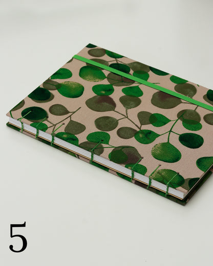 Quaderno per calligrafia con rilegatura copta, copertina in tessuto con foglie di eucalipto verdi su fondo tortora chiaro, elastico e filo verde, occhielli grigi.