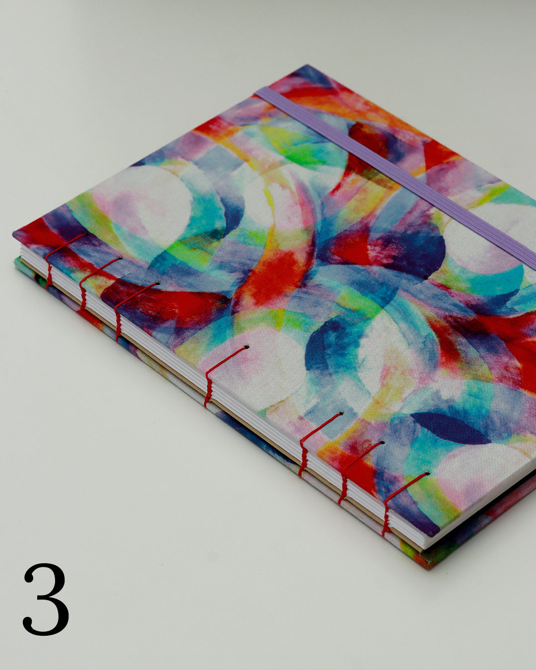 Quaderno per calligrafia con rilegatura copta, copertina in tessuto con cerchi e forme sovrapposte in acquerello multicolore, elastico lilla, filo rosso