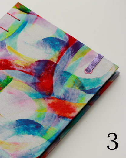 Quaderno per calligrafia con rilegatura copta, copertina in tessuto con cerchi e forme sovrapposte in acquerello multicolore, elastico lilla, filo rosso